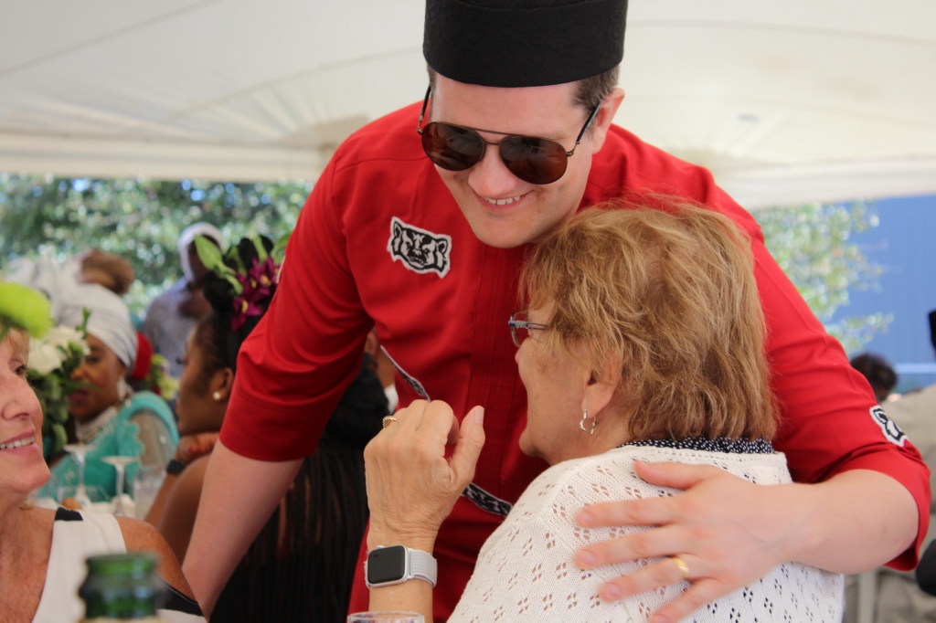 The white groom in an Igbo hat and outfit. The outfit is made from non-traditional material—red with a Wisconsin Badgers logo repeated in a pattern. He is bending over hugging his aunt. 